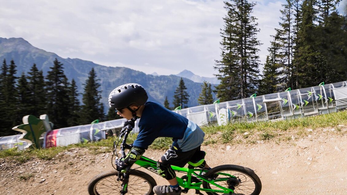 Ein Junge fährt mit einem grünen Fahrrad auf einem staubigen Pfad. Im Hintergrund sind Bäume und Berge zu sehen. | © Roland Haschka