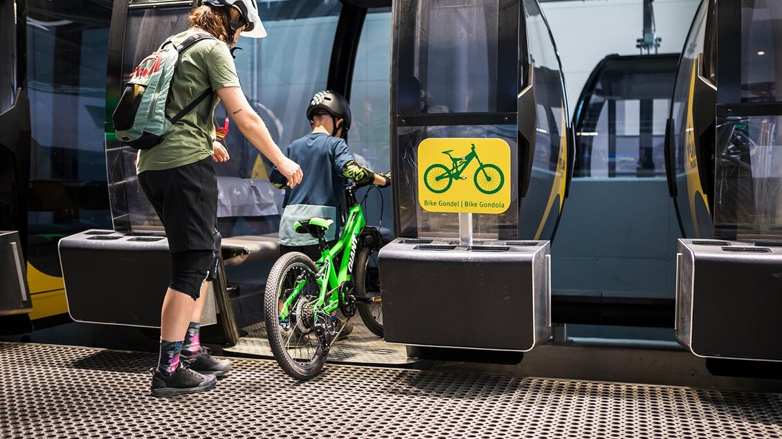 Eine Frau hilft einem Kind, ein grünes Fahrrad in eine Gondel einzuladen. Im Hintergrund sind weitere Gondeln sichtbar. | © Roland Haschka