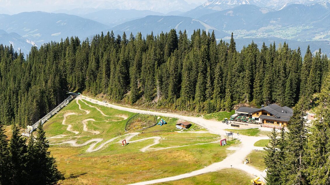 Eine malerische Berglandschaft mit dichten Nadelbäumen und Weiden. Im Vordergrund sind Wege und eine Berghütte sichtbar, umgeben von einer atemberaubenden Kulisse. | © Josh Absenger