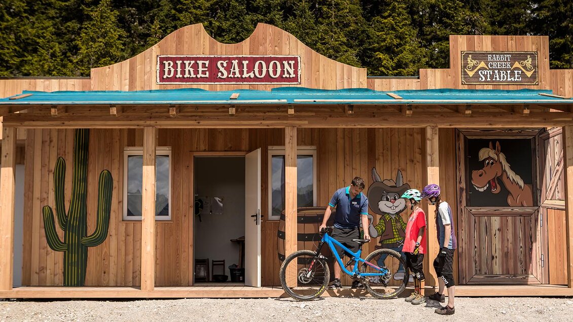 Ein Holzgebäude mit dem Schriftzug "Bike Saloon" und einem freundlichen Empfang. Zwei Radfahrer und ein Mitarbeiter stehen vor dem Eingang und unterhalten sich. | © Roland Haschka