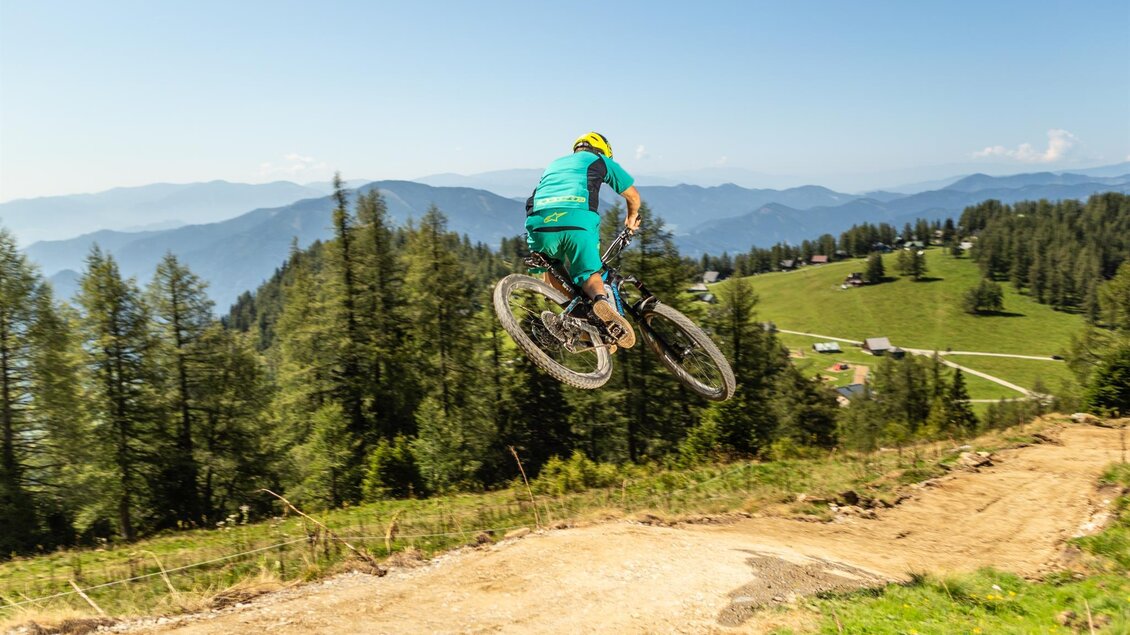 Ein Radfahrer springt mit seinem Mountainbike über einen Hügel in der Natur. Im Hintergrund sind Wälder und Berge zu sehen.