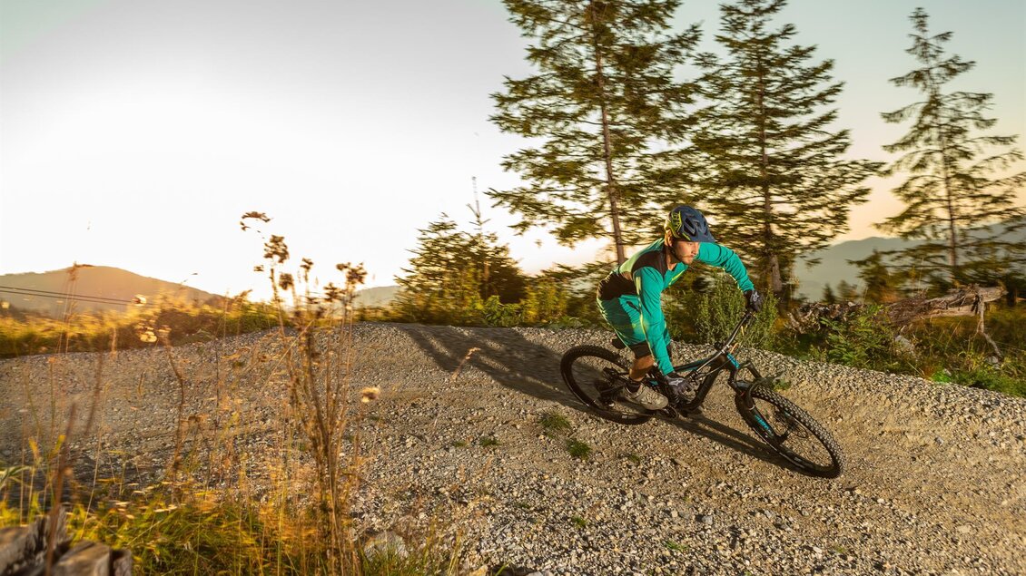 Ein Mountainbiker fährt schnell auf einem schottrigen Weg. Im Hintergrund sind Bäume und ein klarer Himmel zu sehen.