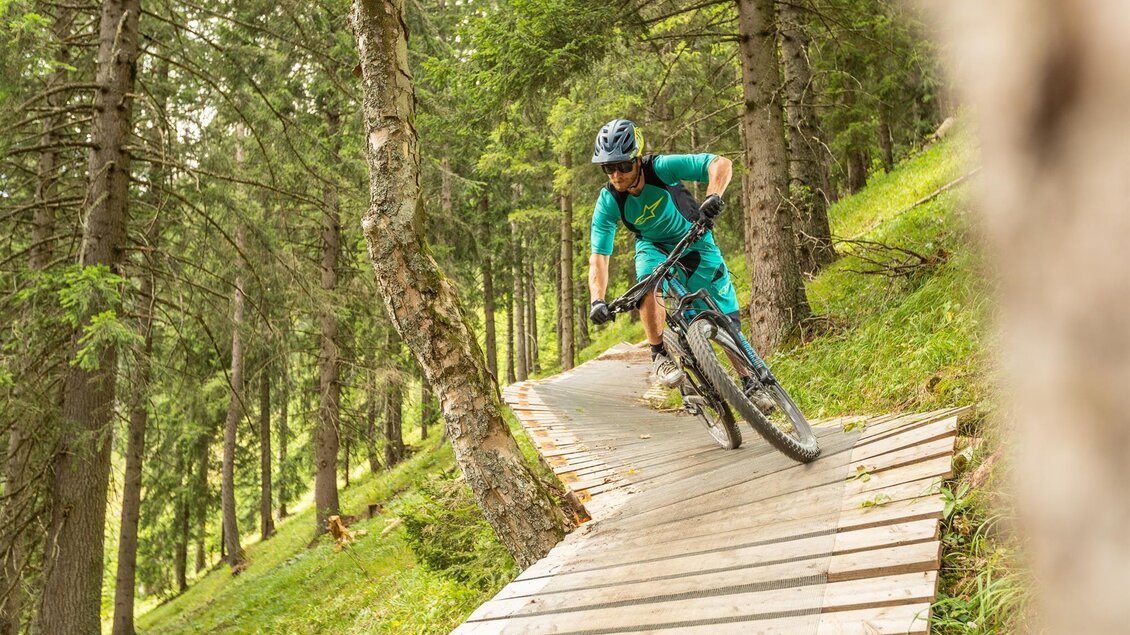 Ein Mountainbiker fährt auf einem Holztrail durch einen Wald. Die Umgebung ist grün und sonnig.
