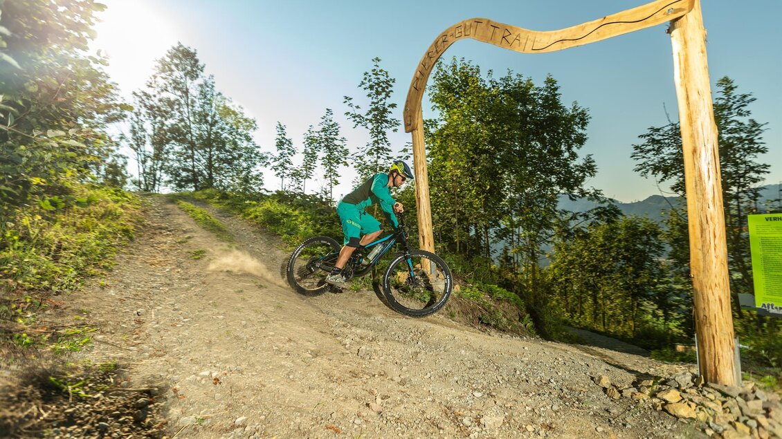 Ein Radfahrer in blaugrüner Ausrüstung fährt auf einem Schotterweg. Im Hintergrund ist ein Tor aus Holz und eine grüne Landschaft zu sehen.