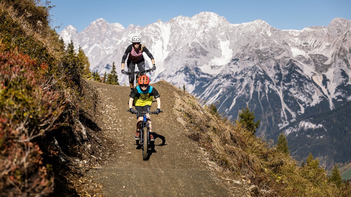 Zwei Radfahrer fahren einen Bergweg entlang, umgeben von majestätischen Bergen. Der Himmel ist klar und die Landschaft ist sonnig und einladend. | © Roland Haschka | Alpreif