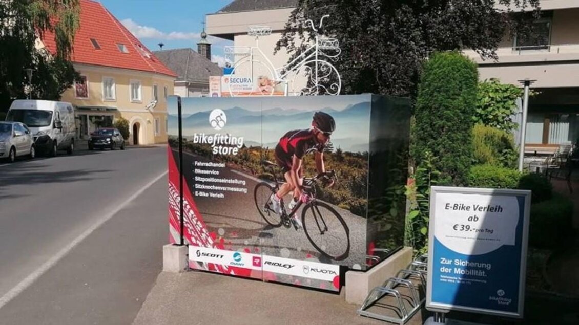 Ein Werbeschild für einen Fahrradladen zeigt einen Radfahrer in action. Im Vordergrund steht auch ein Schild zur E-Bike-Verleihung. | © Bikefitting Store_Hiebler