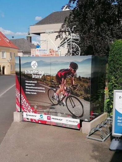An advertisement sign for a bicycle shop shows a cyclist in action. In the foreground, there is also a sign for e-bike rental. | © Bikefitting Store_Hiebler