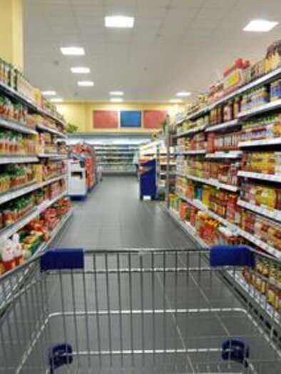 A shopping cart is in the aisle of a supermarket. The shelves are filled with various foods and drinks. | © Symbolfoto Fotolia