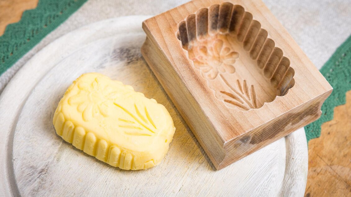 Eine handgefertigte Seife in Form einer Blume liegt neben einer hölzernen Form. Die Seife hat eine gelbe Farbe und die Form zeigt ein florales Muster. | © Netzwerk Kulinarik wildbild