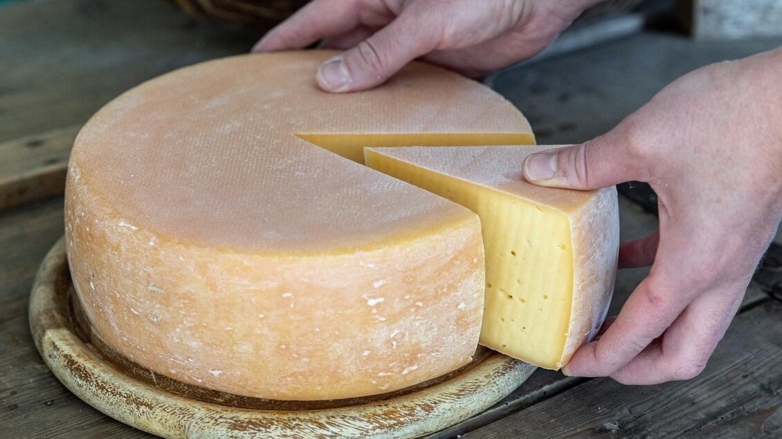 Ein runder Käse wird in Stücke geschnitten. Eine Hand hält ein Stück Käse, während das andere Stück bereits herausgenommen wurde. | © Martin Huber