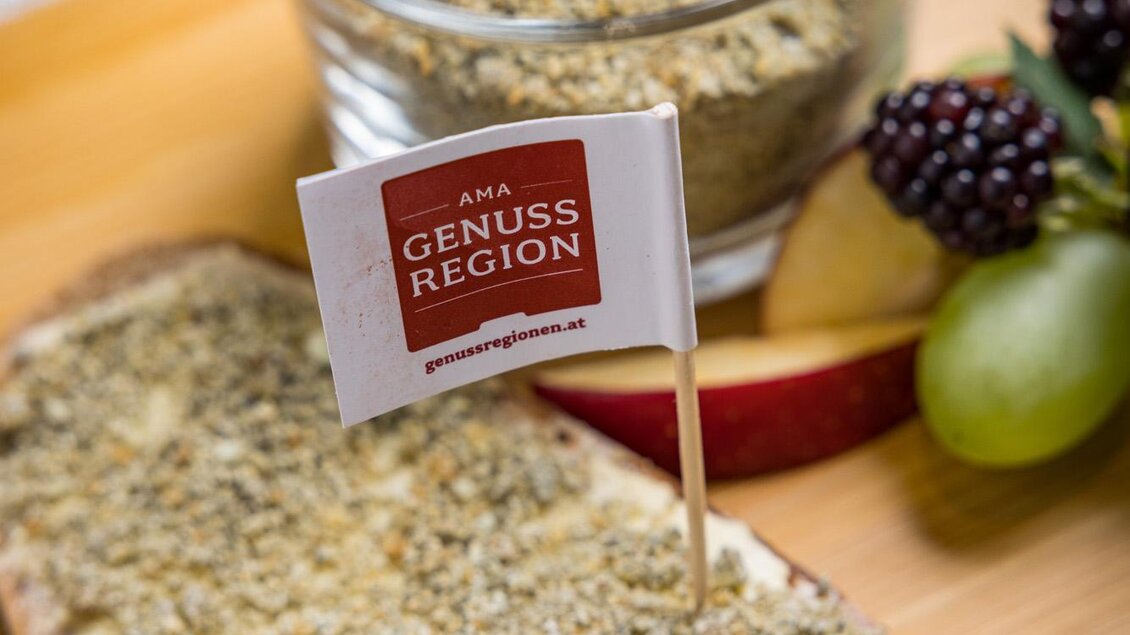 Ein Stück Brot mit einer würzigen Belag, garniert mit einer kleinen Flagge "Genuss Region". Im Hintergrund befindet sich ein Glas mit ähnlichem Belag sowie einige frische Früchte. | © Netzwerk Kulinarik wildbild