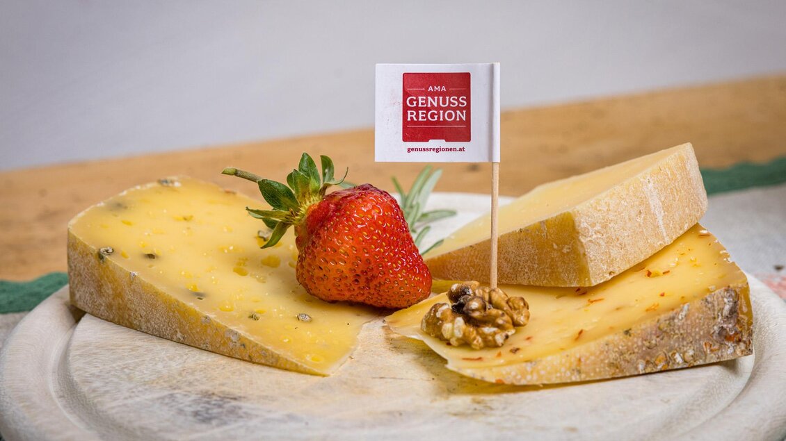 Ein rustikaler Holzbrett mit verschiedenen Käsesorten, einer Erdbeere und einer Walnuss. Ein kleiner Fahne mit der Aufschrift "Genussregion" steht in der Mitte. | © Netzwerk Kulinarik wildbild