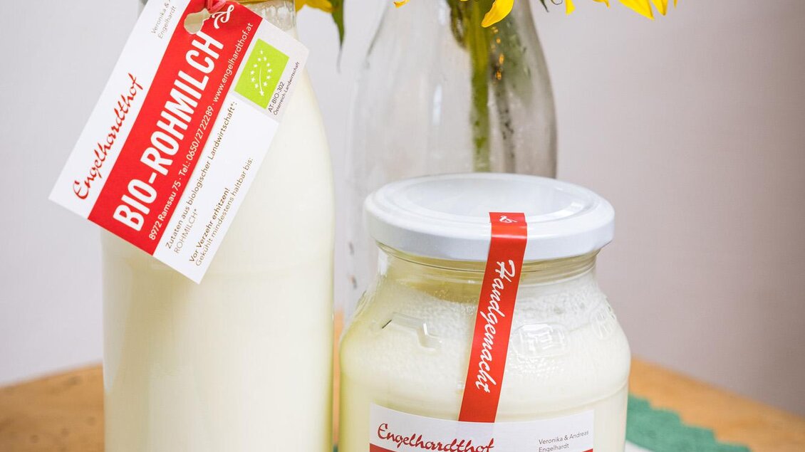 Eine Flasche und ein Glas mit Bio-Milchprodukten stehen auf einem Tisch, umgeben von einer Vase mit Sonnenblumen. Der Hintergrund ist eine einfache Tischdecke mit Blumenmuster. | © Netzwerk Kulinarik wildbild