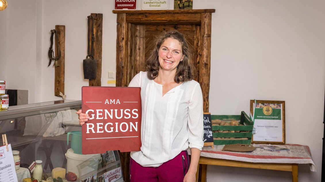 Eine lächelnde Frau hält ein Schild mit der Aufschrift "AMA GENUSS REGION". Im Hintergrund befinden sich Produkte in einem Lebensmittelgeschäft. | © Netzwerk Kulinarik wildbild