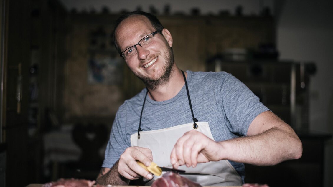 Ein lächelnder Mann in einer Küche schneidet Fleisch mit einem Messer. Auf dem Tisch liegen weitere Fleischstücke. | © Stefan Leitner