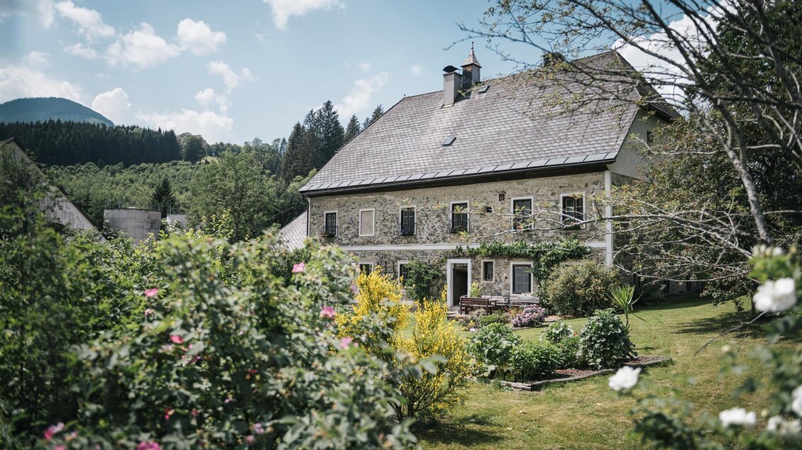 Ein charmantes Haus umgeben von einem grünen Garten und bunten Blumen. Der Himmel ist klar und die Landschaft ist hügelig. | © Stefan Leitner