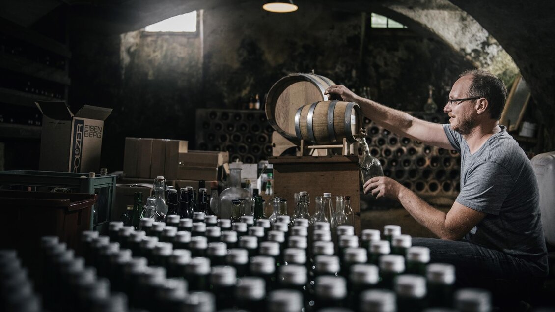 Ein Mann arbeitet in einem Weinkeller und füllt Flüssigkeit aus einem Fass in Flaschen. Im Hintergrund sind viele Flaschen und Kisten zu sehen. | © Stefan Leitner