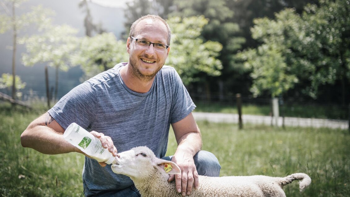 Ein Mann füttert ein Lamm mit einer Flasche. Im Hintergrund sind Bäume und eine ländliche Landschaft sichtbar. | © Stefan Leitner