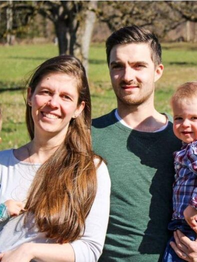 A happy family stands in a green field. The parents are holding their two small children in their arms and radiate joy. | © Lienlbauer