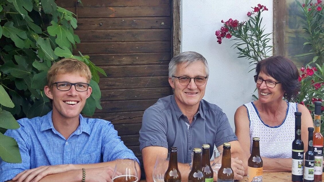 Eine Gruppe von drei Menschen sitzt an einem Holztisch im Freien. Verschiedene Getränke und Lebensmittel sind auf dem Tisch präsentiert. | © Bio-Obsthof Kirchengast