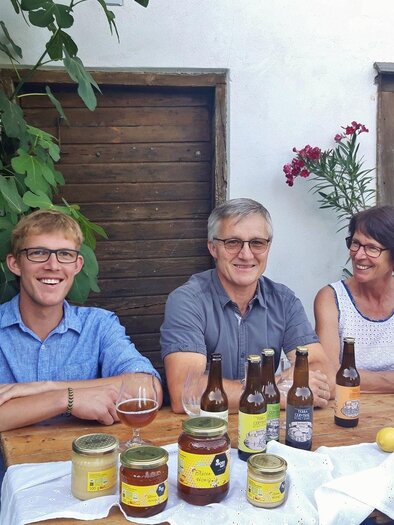Eine Gruppe von drei Menschen sitzt an einem Holztisch im Freien. Verschiedene Getränke und Lebensmittel sind auf dem Tisch präsentiert. | © Bio-Obsthof Kirchengast