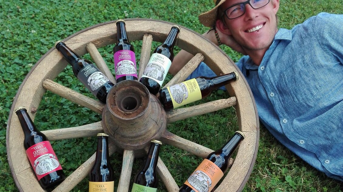 Ein Mann liegt lächelnd neben einem Holzrad, das mit verschiedenen Flaschen umrandet ist. Die Flaschen enthalten unterschiedliche Getränke und sind farbenfroh gestaltet. | © Bio-Obsthof Kirchengast