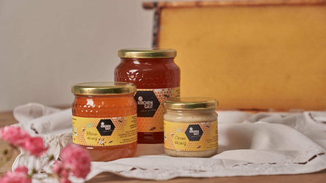 Drei Gläser mit Honig in verschiedenen Größen stehen auf einem Tisch. Im Hintergrund liegt eine Bienenwabe, und einige Blumen vervollständigen die sanfte Atmosphäre. | © Philipp Monohart