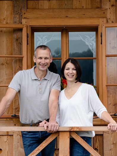 A friendly couple stands on the porch of a wooden house. They are smiling and appear happy.