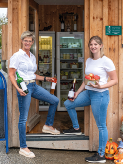 Zwei Frauen stehen an einem kleinen Verkaufsstand und halten Flaschen und ein Körbchen mit Äpfeln. Neben ihnen steht ein Schild mit der Aufschrift "BIO Äpfel". | © Familie Lammer