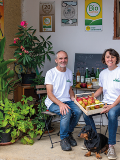 A couple is sitting in front of a building with plants and wooden crates. They are holding a basket of apples and a small dog is sitting next to them. | © Familie Meyer