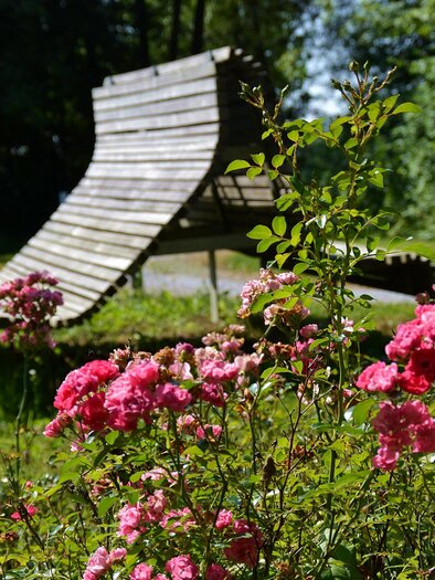 Eine gemütliche Liege aus Holz steht in einem grünen Garten. Im Vordergrund blühen rosa Rosen. | © Kurkommission Bad Blumau