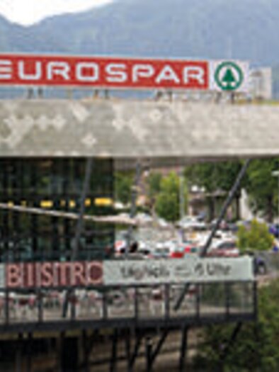 A Eurospar supermarket with a modern entrance area. In the foreground, there is a bistro with an outdoor seating area. | © C.Stein
