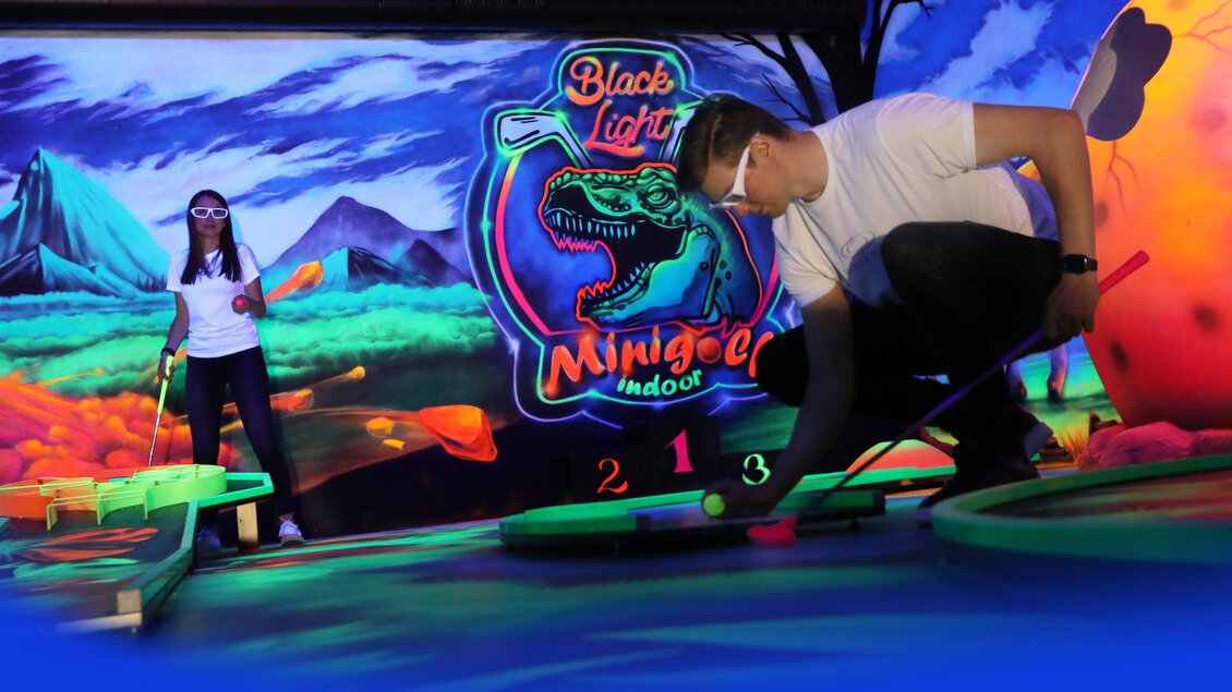 Ein aufregendes Minigolfspiel im Schwarzlicht mit leuchtenden Farben und einer Dinosaurier-Themenwand. Zwei Personen spielen, während die bunten Elemente im Hintergrund strahlen. | © Black Light Minigolf