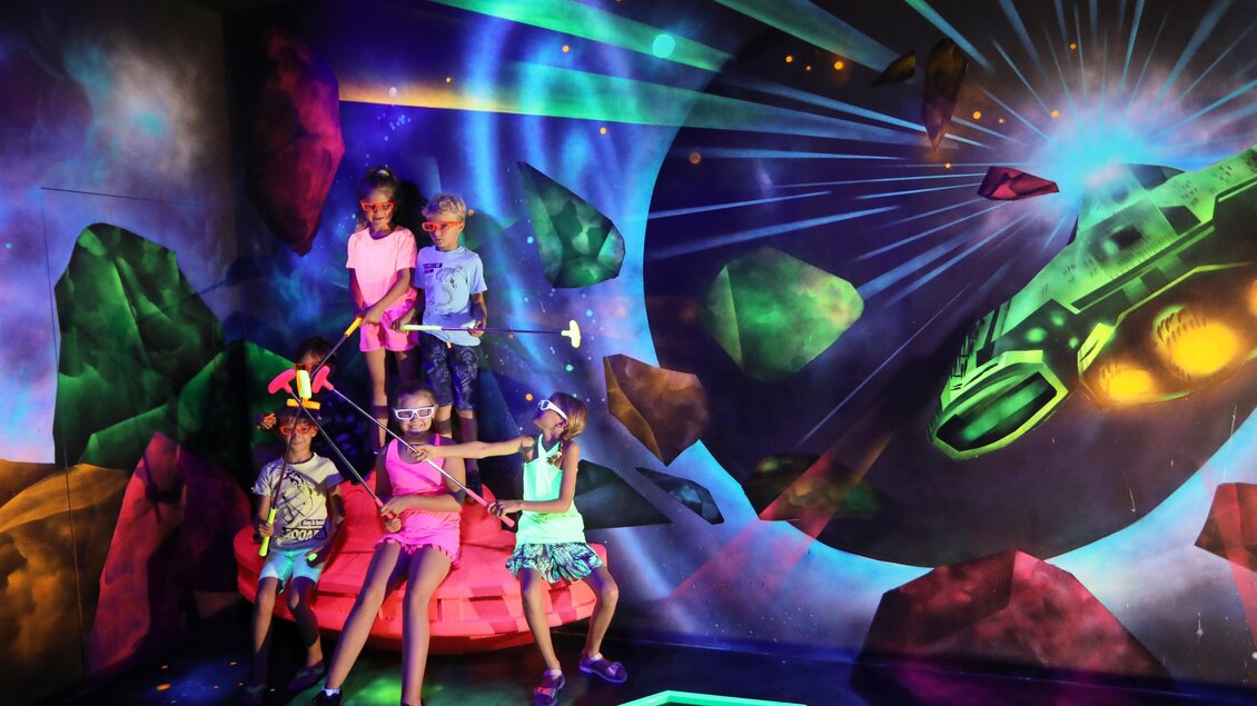 Eine Gruppe von Kindern steht in einem bunten Raum mit Weltraummotiv. Sie posieren begeistert vor einer riesigen Wandmalerei eines Raumschiffs und farbenfrohen Asteroiden. | © Black Light Minigolf