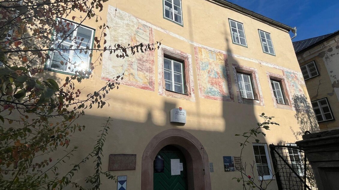 Ein historisches Gebäude mit bunten Wandmalereien und grüner Eingangstür. Die Fenster reflektieren das Sonnenlicht und die Umgebung ist von Pflanzen umgeben. | © TVB Ausseerland Salzkammergut_Nathalie Schröttenhamer