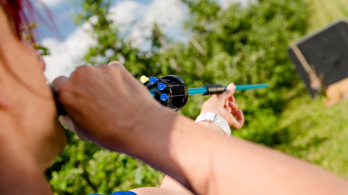 Eine Person zielt mit einem Pfeil und Bogen in einer grünen Umgebung. Im Hintergrund ist ein Ziel sichtbar. | © Mias Photoart