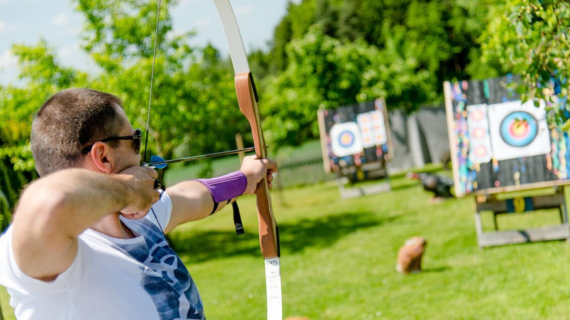 Eine Person beim Bogenschießen in einem grünen Garten. Im Hintergrund sind Zielscheiben zu sehen. | © Mias Photoart