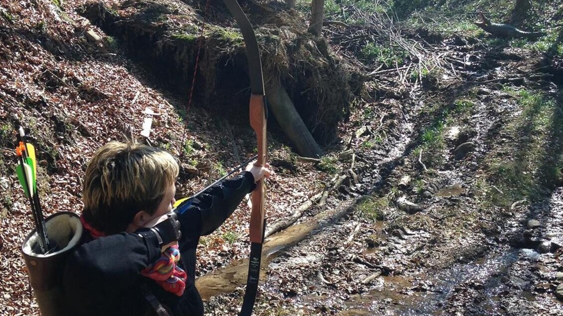 Ein Junge steht im Wald und zielt mit einem Bogen. Um ihn herum sind Blätter und ein kleiner Bach. | © Bogenparcours Siebernegg