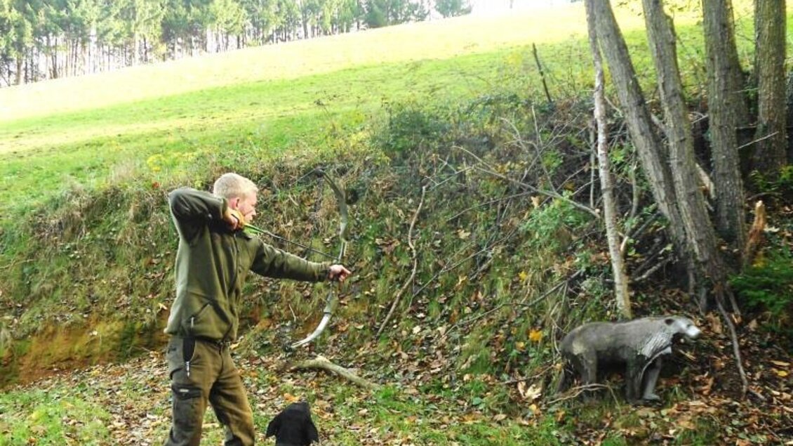 Ein Mann wirft einen Stock, während ein schwarzer Hund daneben steht. Im Hintergrund ist ein Wildschwein zu sehen, umgeben von Bäumen und einer grünen Wiese. | © Bogenparcours Siebernegg