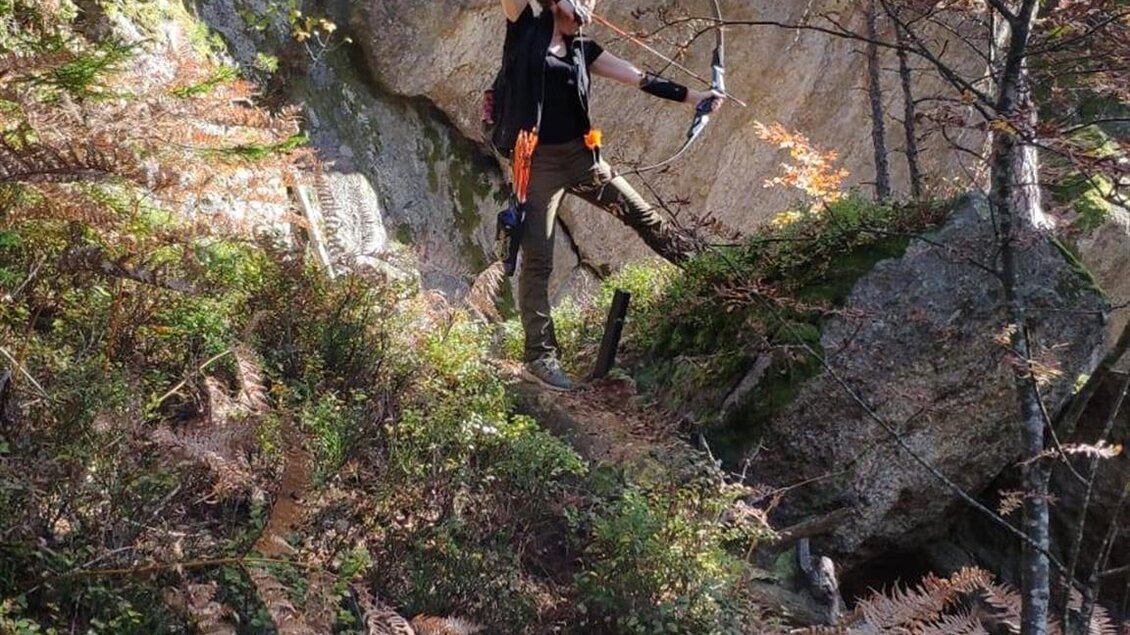 Eine Person steht auf einem steilen Hang und richtet einen Bogen auf ein Ziel. Im Hintergrund sind Felsen und Bäume zu sehen. | © Bogenparcours Siebernegg