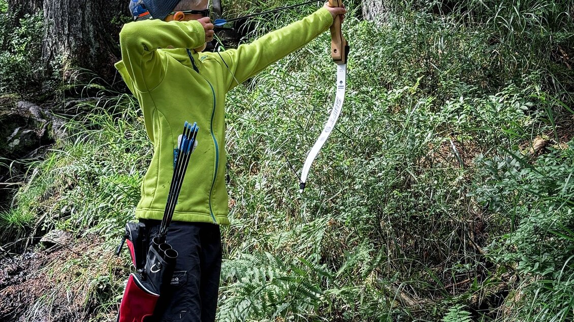 Ein Junge schießt mit einem Bogen in einem Wald. Er trägt eine grüne Jacke und steht auf einem schmalen Weg zwischen Bäumen. | © Daniel Radauer