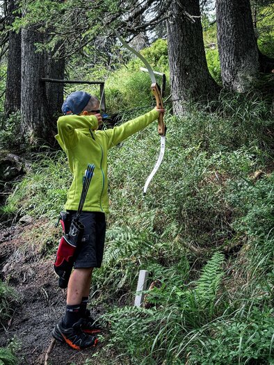 A boy is shooting with a bow in a forest. He is wearing a green jacket and standing on a narrow path between trees. | © Daniel Radauer