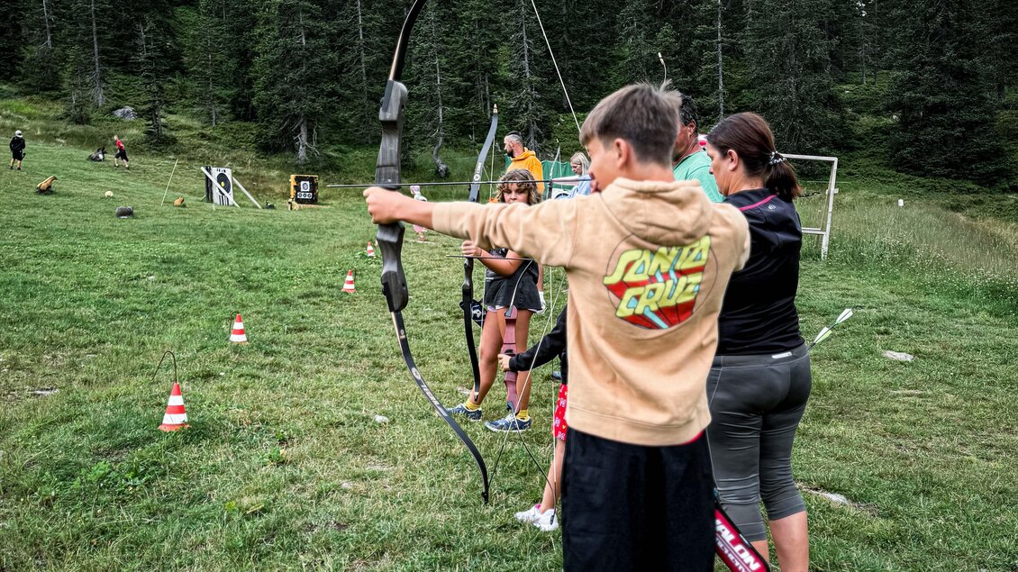 Ein Junge zielt mit einem Bogen auf ein Ziel in einer bewaldeten Umgebung. Im Hintergrund sind weitere Personen zu sehen, die ebenfalls am Bogenschießen teilnehmen. | © Daniel Radauer