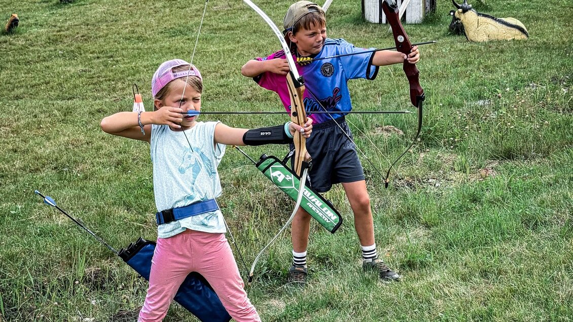 Zwei Kinder üben das Bogenschießen auf einer Wiese. Im Hintergrund sind Zielscheiben sichtbar und weitere Kinder in Aktion. | © Daniel Radauer