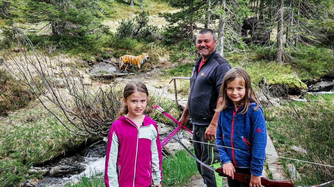 Eine Gruppe von drei Personen steht im Wald neben einem Bach. Zwei Mädchen halten Pfeile, während ein Erwachsener sie an einem Ausflugsort begleitet. | © Daniel Radauer