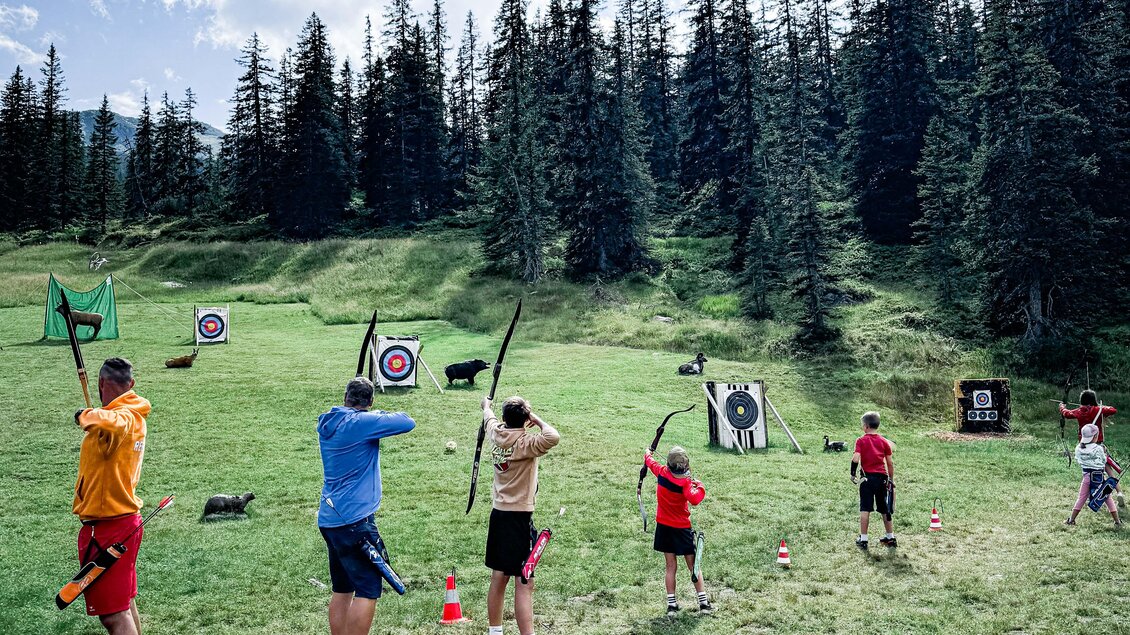 Eine Gruppe von Menschen übt das Bogenschießen in einer grünen Wiese. Im Hintergrund sind Zielscheiben und hohe Tannenbäume zu sehen. | © Daniel Radauer