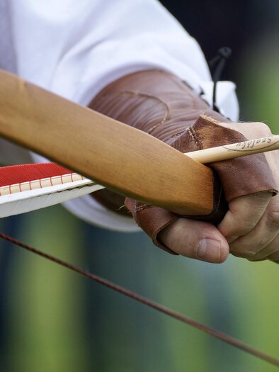 Ein Bogen wird gespannt, mit einem Pfeil in der Hand des Schützen. Der Hintergrund zeigt eine grüne Wiese. | © Symbolfoto Pixabay