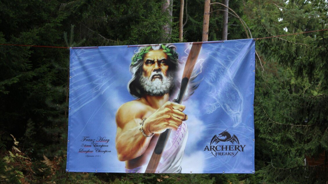 Ein großes Banner zeigt eine mythologische Figur mit einem Bogen in der Hand. Die Darstellung ist umgeben von grünen Bäumen im Hintergrund. | © Archery Freaks - Frahs