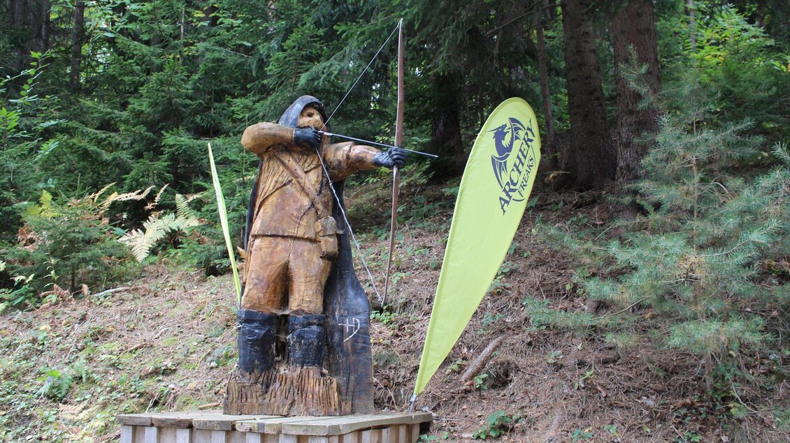 Eine Holzskulptur eines Bogenschützen steht im Wald. Daneben weht ein grünes Schild mit einem Logo. | © Archery Freaks - Frahs