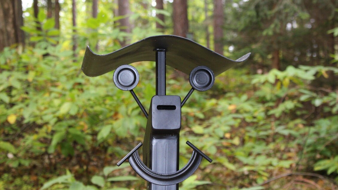 Ein modernes Kunstwerk in Form eines Gesichts aus Metall, umgeben von Bäumen und grünen Pflanzen. Die einfachen Formen und Farben erzeugen eine freundliche Stimmung. | © Archery Freaks - Frahs